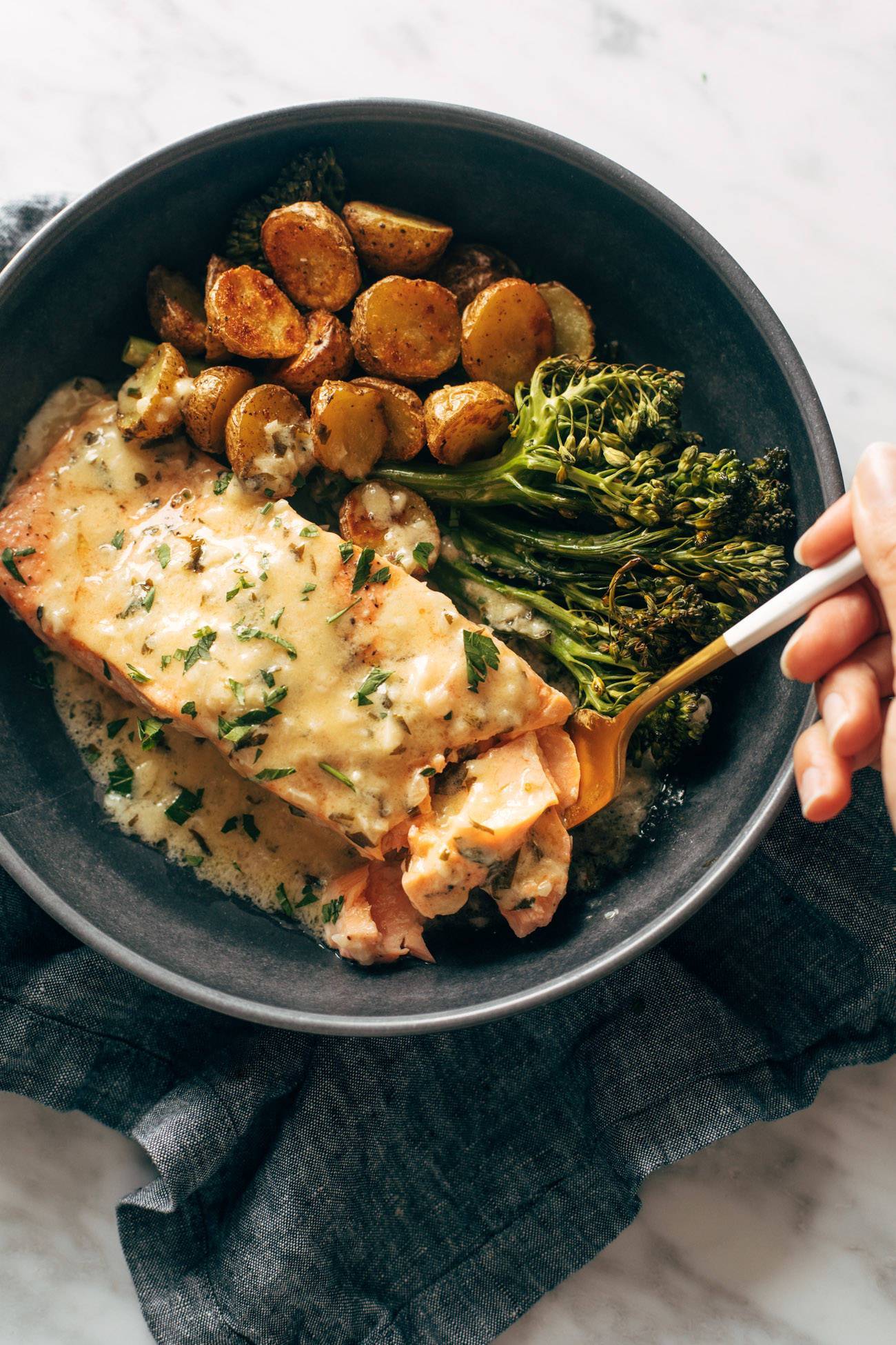 White hand holding a fork and grabbing a bite of baked salmon. Baked salmon is in a bowl with broccoli and roasted potatoes and is smothered in lemon sauce.